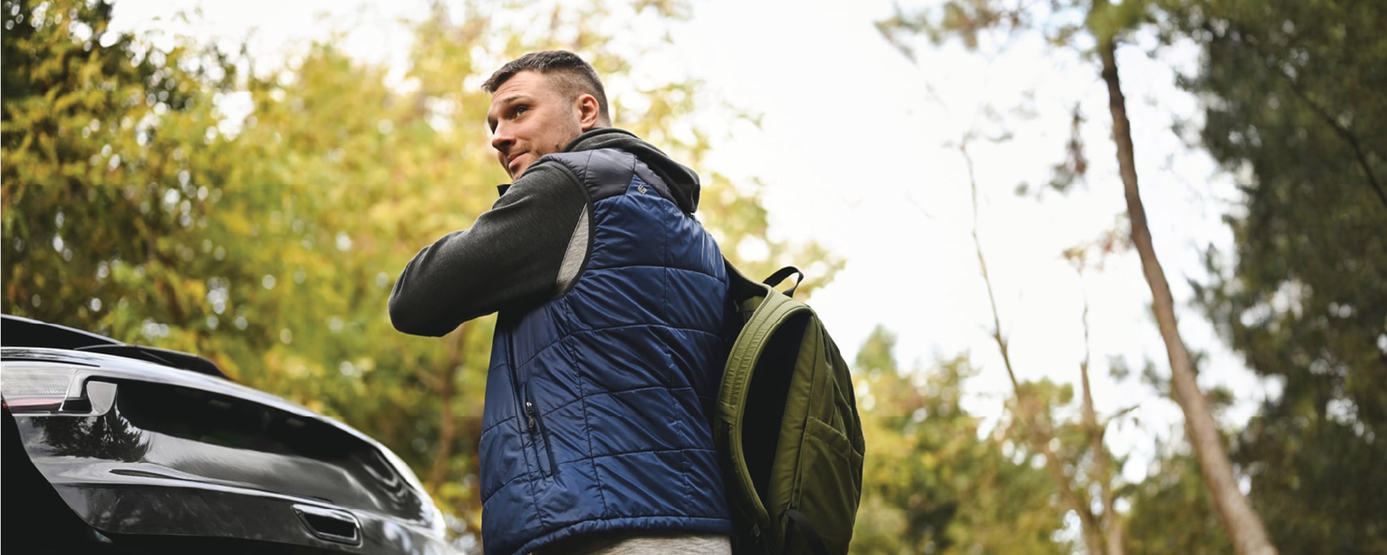 Person wearing a blue quilted vest over a dark hoodie adjusts a backpack while standing beside the open trunk of a car in a wooded area, looking back over their shoulder.