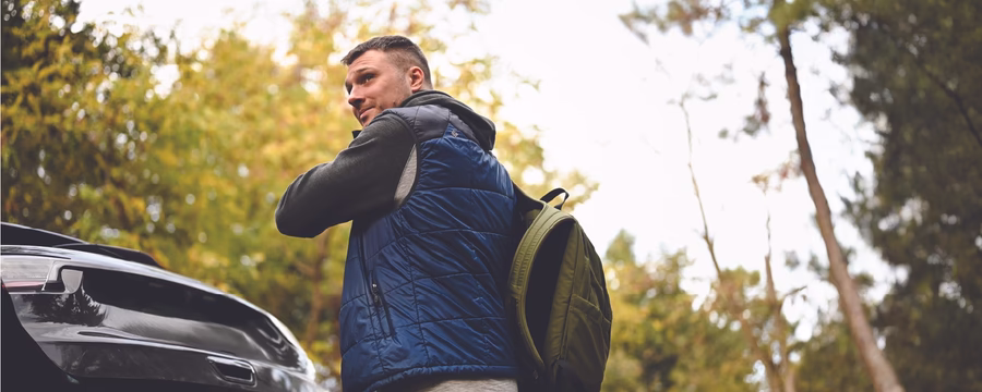 Person wearing a blue quilted vest over a dark hoodie adjusts a backpack while standing beside the open trunk of a car in a wooded area, looking back over their shoulder.