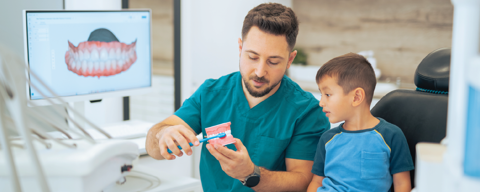 A dental professional in teal scrubs demonstrates proper toothbrushing technique using a large model of teeth and a toothbrush while a young boy watches attentively in a dental exam room. A monitor displaying a dental image is visible in the background.