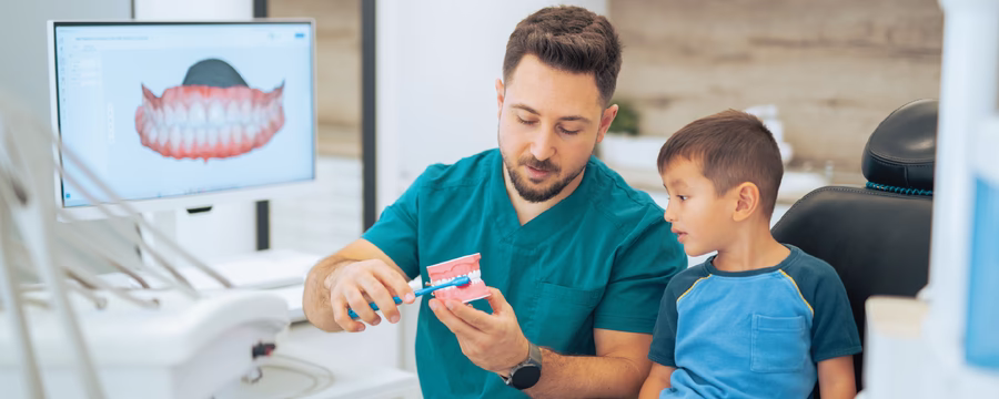 A dental professional in teal scrubs demonstrates proper toothbrushing technique using a large model of teeth and a toothbrush while a young boy watches attentively in a dental exam room. A monitor displaying a dental image is visible in the background.