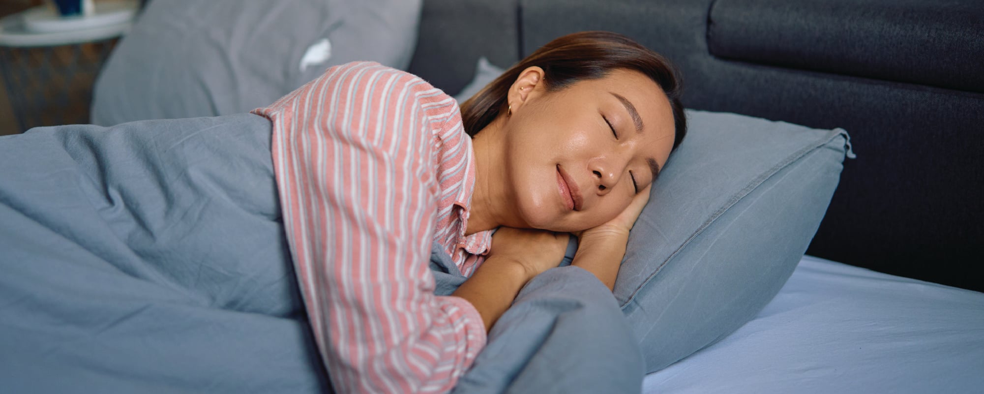 A person sleeps peacefully on their side in bed, wearing a pink-and-white striped pajama top and resting on a light blue pillow under a matching blue comforter, with a dark upholstered headboard in the background.