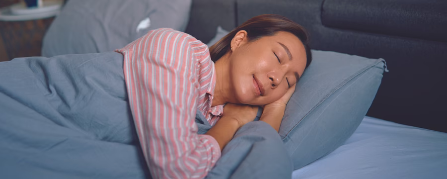 A person sleeps peacefully on their side in bed, wearing a pink-and-white striped pajama top and resting on a light blue pillow under a matching blue comforter, with a dark upholstered headboard in the background.