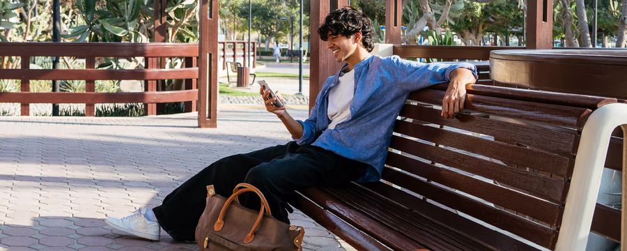 Young man sitting on a park bench, smiling while looking at his smartphone with a travel bag beside him.