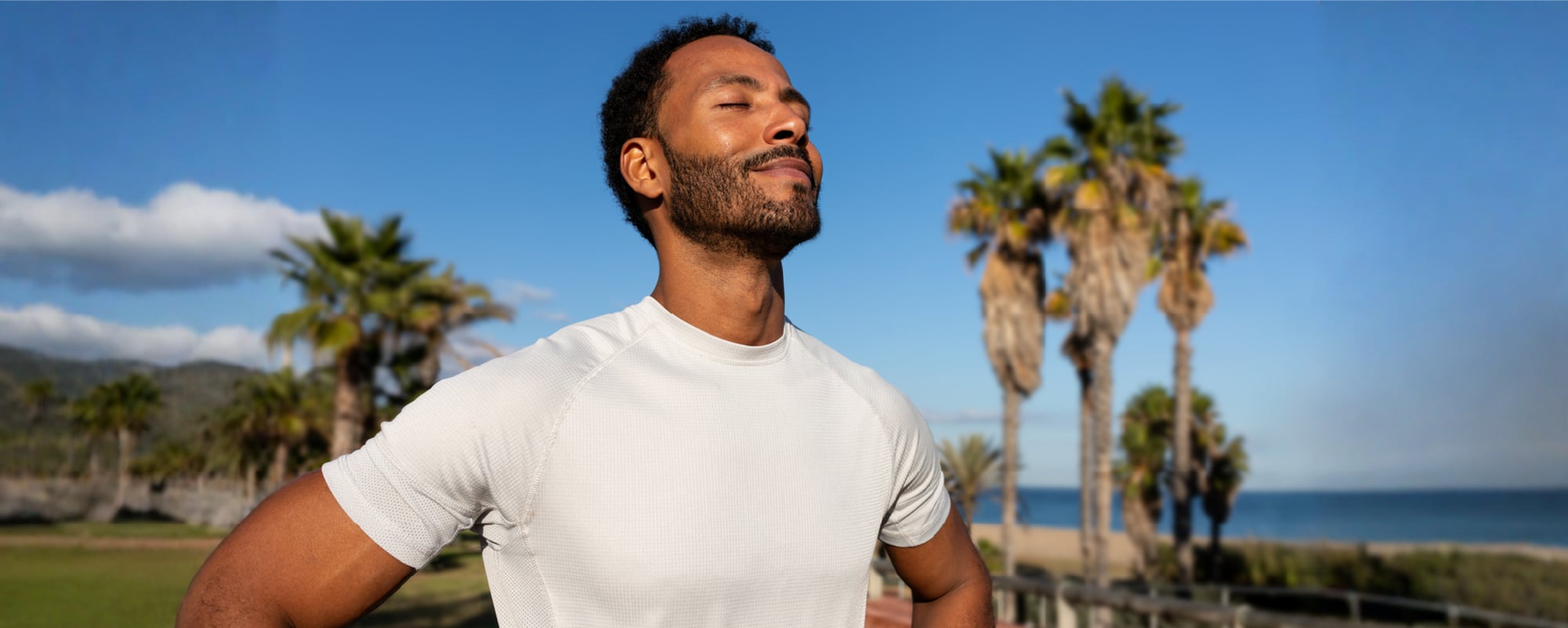 Athletic man pausing after a run near a palm-lined beach with his eyes closed.