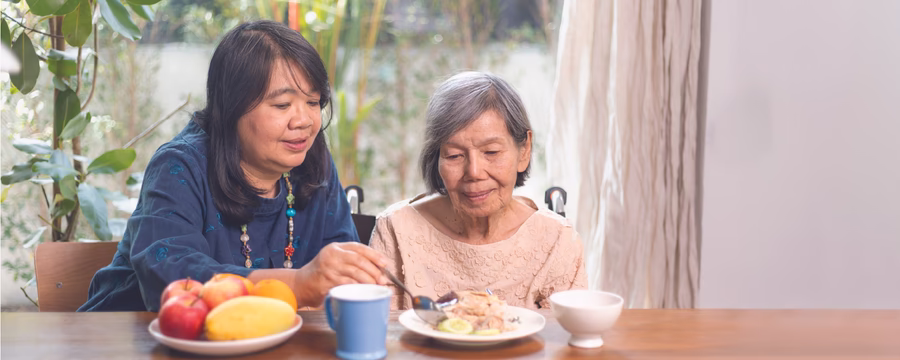 A woman assists an elderly woman at a table, helping her eat a meal in a bright room with greenery visible outside.