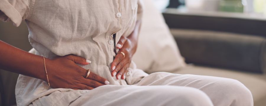 Person sitting on a bed in light-colored clothing, holding their lower abdomen with both hands, suggesting stomach pain, cramps, or abdominal discomfort. The background is softly blurred, creating a calm indoor setting.