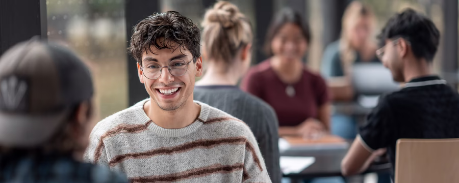 Smiling young man wearing glasses and a striped sweater talks with another student in a busy college classroom.