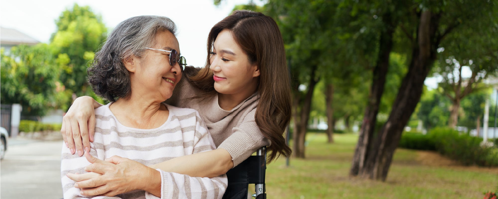 Smiling woman embraces an older woman seated in a wheelchair at a park.
