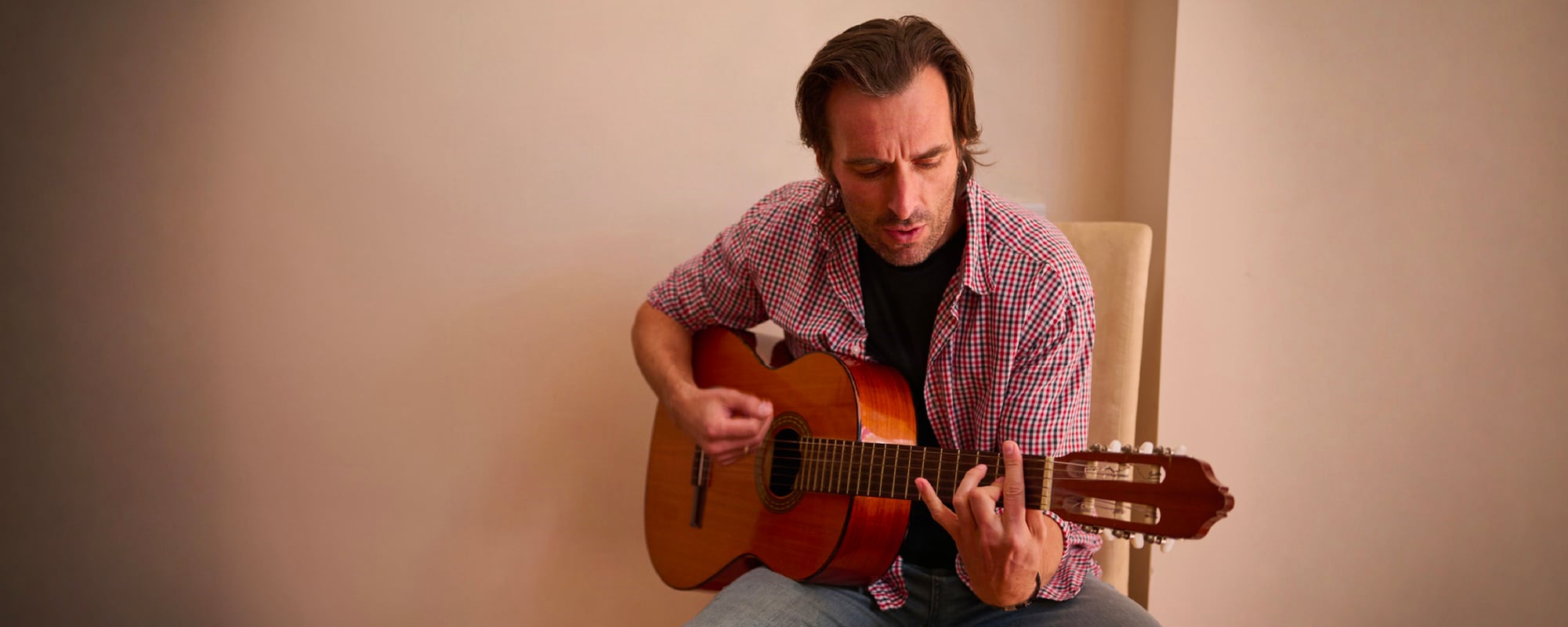 Man seated indoors playing an acoustic guitar, wearing a red plaid shirt over a black T-shirt.