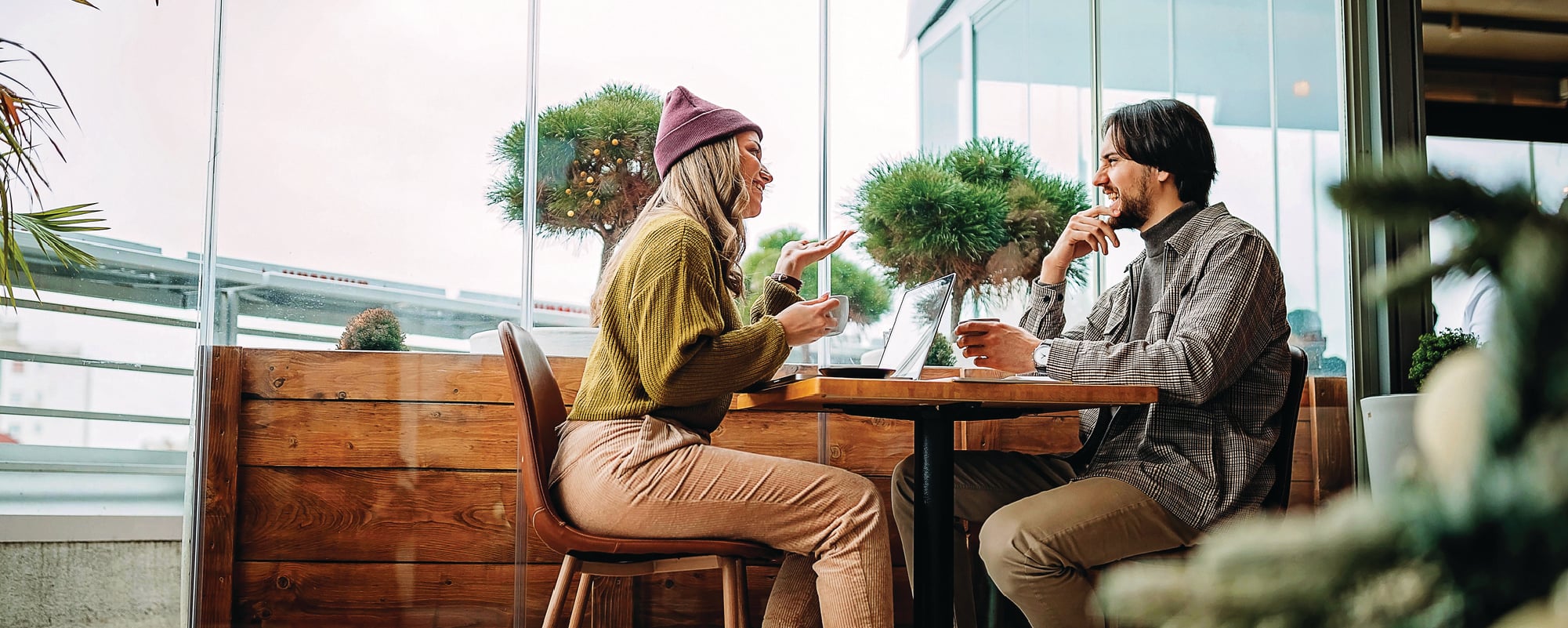 Two people sit across from each other at a café table, smiling and chatting over coffee. One gestures while speaking, and an open laptop sits between them near large windows with potted greenery in the background.