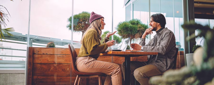 Two people sit across from each other at a café table, smiling and chatting over coffee. One gestures while speaking, and an open laptop sits between them near large windows with potted greenery in the background.