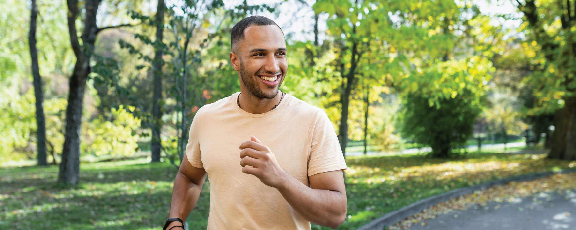 A smiling man jogs along a paved path in a green park, surrounded by trees and sunlight on a clear day.