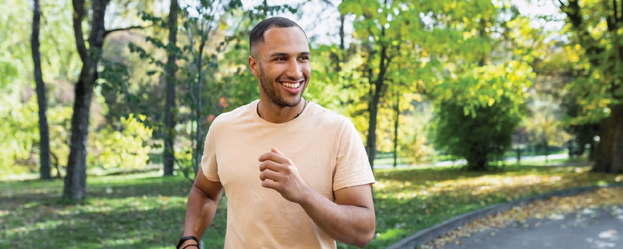 A smiling man jogs along a paved path in a green park, surrounded by trees and sunlight on a clear day.