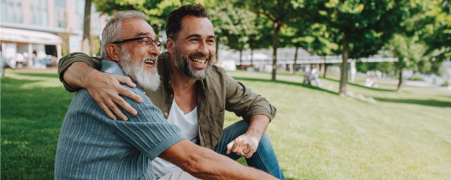 An older man with a white beard and glasses and a younger man sit together on the grass in a park, smiling and embracing as they enjoy time outdoors on a sunny day.