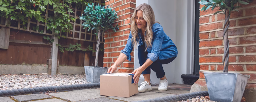 Smiling person crouching outside a front door, placing or picking up a cardboard package on the doorstep. The entryway is framed by brick walls and potted plants, suggesting a home delivery scene.