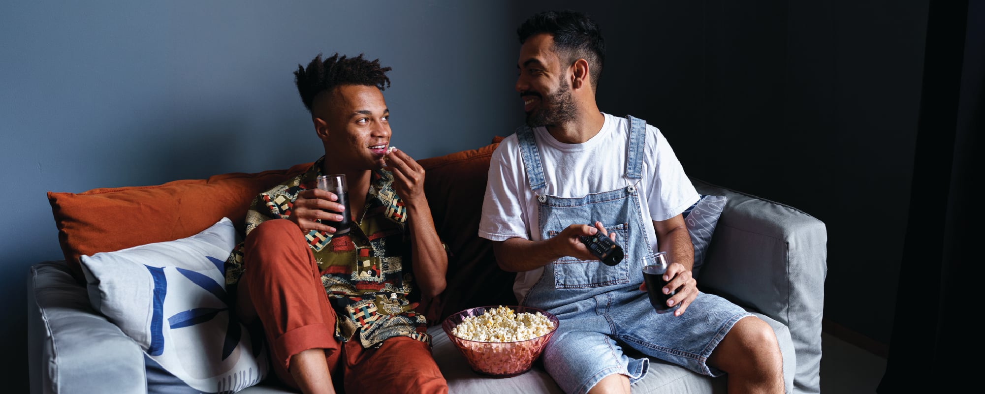 Two people sit together on a couch watching something off-camera, sharing popcorn and drinks. One person is holding a game controller while smiling at the other, creating a relaxed, social movie-or-game-night atmosphere. A bowl of popcorn rests between them on the sofa.