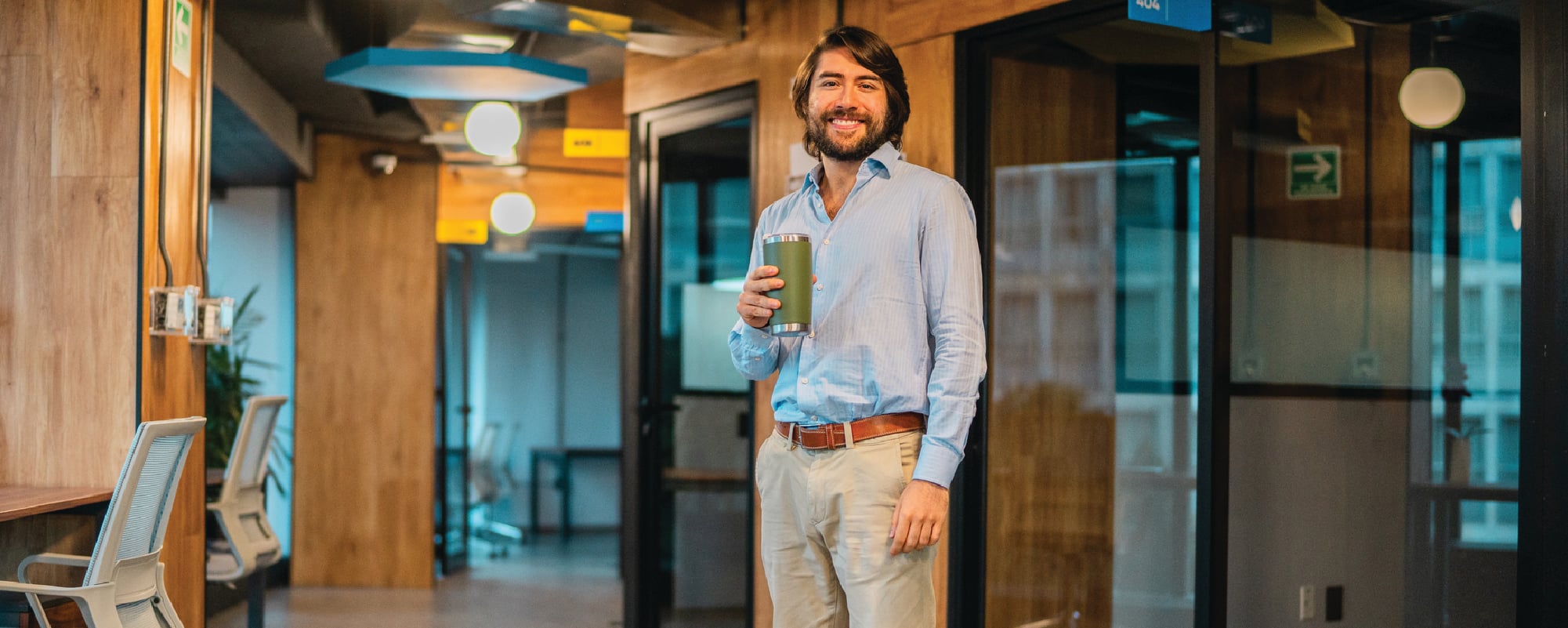A smiling man stands in a modern office hallway holding a reusable travel tumbler. He is wearing a light blue button-up shirt and khaki pants, with glass-walled meeting rooms and warm wood accents visible in the background.