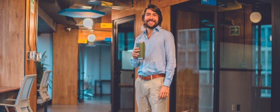 A smiling man stands in a modern office hallway holding a reusable travel tumbler. He is wearing a light blue button-up shirt and khaki pants, with glass-walled meeting rooms and warm wood accents visible in the background.