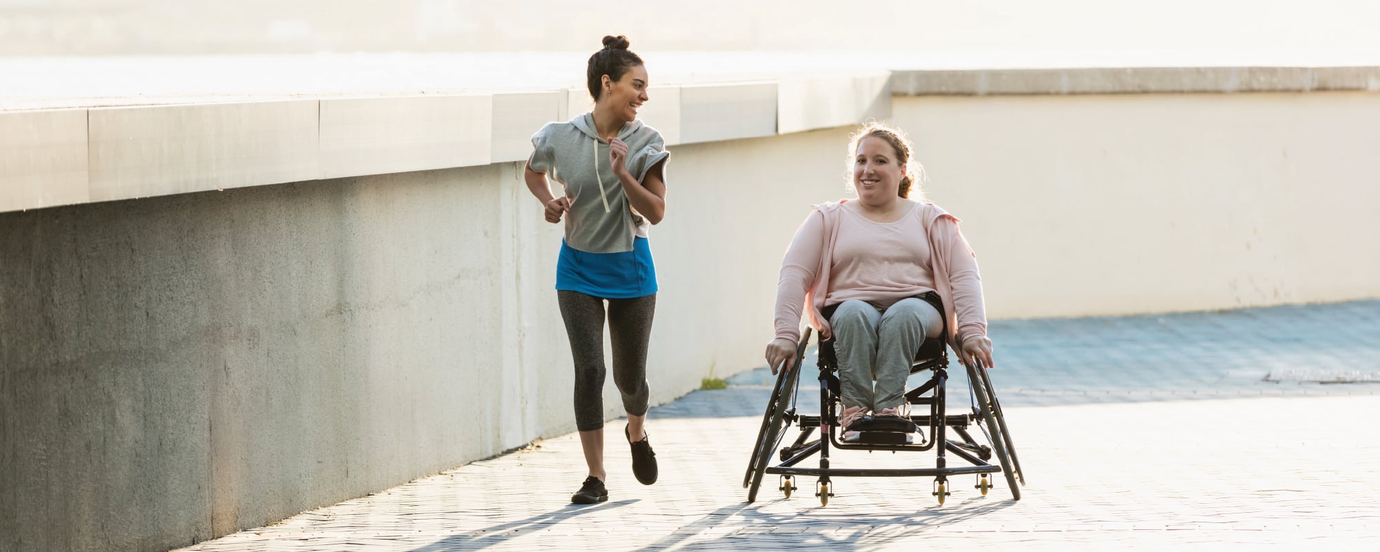 wo women exercise outdoors, one jogging alongside the other as she moves in a racing wheelchair.