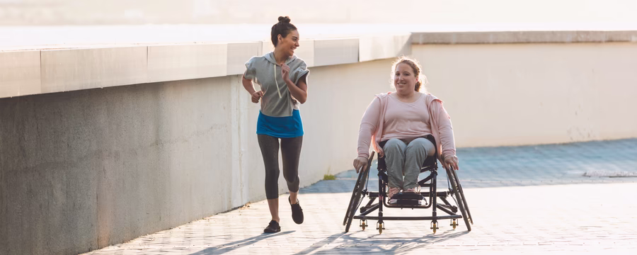 wo women exercise outdoors, one jogging alongside the other as she moves in a racing wheelchair.