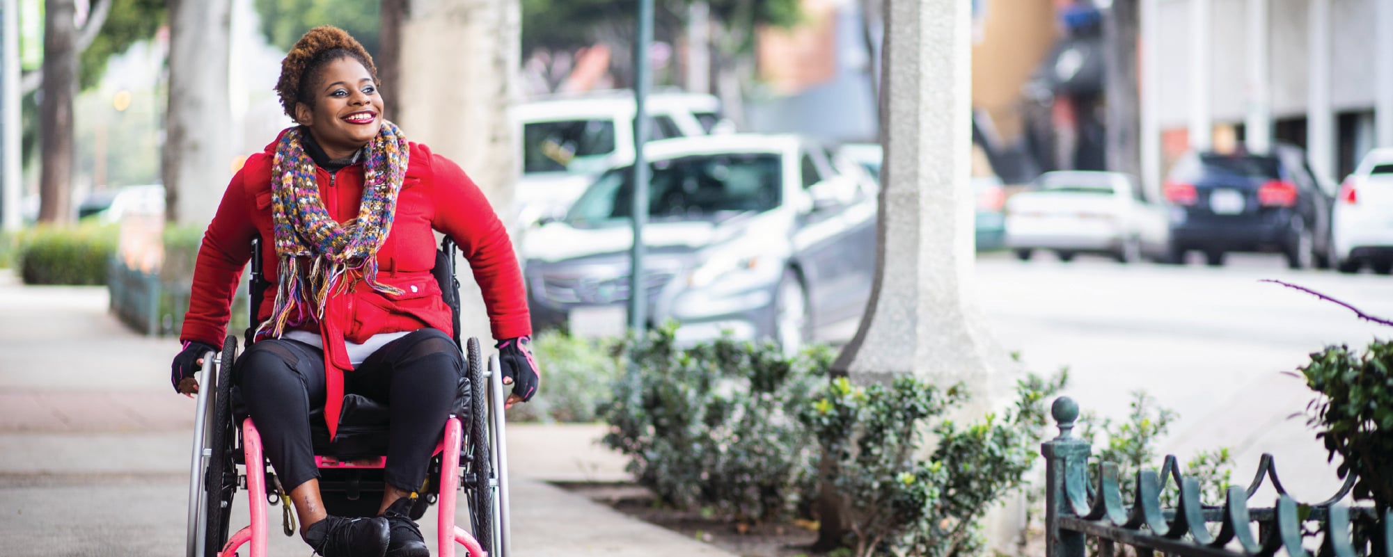 A smiling person using a wheelchair moves along a city sidewalk lined with trees and low shrubs. They are wearing a bright red coat, a colorful patterned scarf, dark pants, and gloves. Parked cars and urban buildings appear blurred in the background.