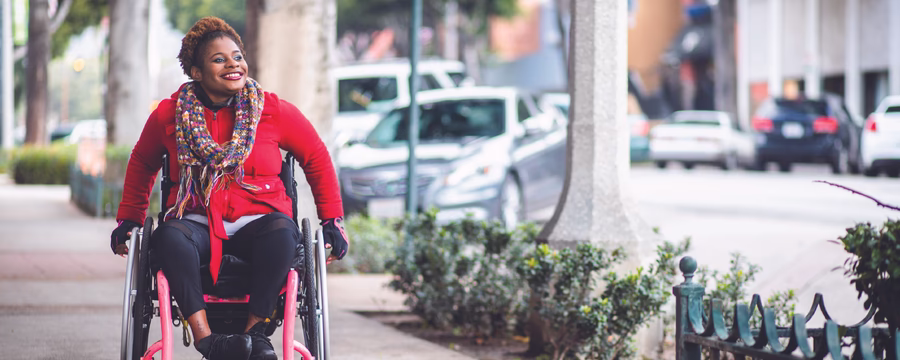 A smiling person using a wheelchair moves along a city sidewalk lined with trees and low shrubs. They are wearing a bright red coat, a colorful patterned scarf, dark pants, and gloves. Parked cars and urban buildings appear blurred in the background.