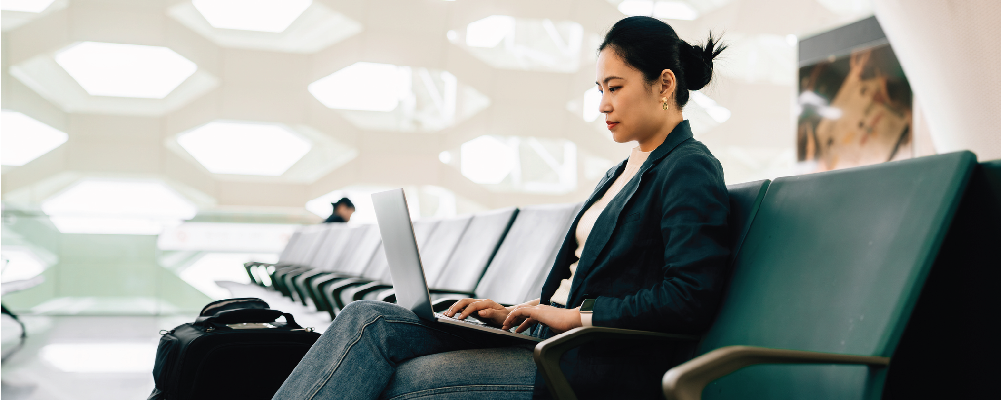 A woman sits in an airport waiting area, working on a laptop while seated on a row of green chairs. A suitcase and bag are beside her, and the bright, modern terminal features geometric wall openings in the background.