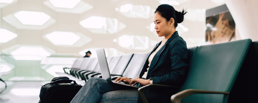 A woman sits in an airport waiting area, working on a laptop while seated on a row of green chairs. A suitcase and bag are beside her, and the bright, modern terminal features geometric wall openings in the background.