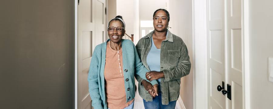 Older woman wearing glasses and a teal cardigan walks down a hallway while a younger woman gently supports her by the arm.