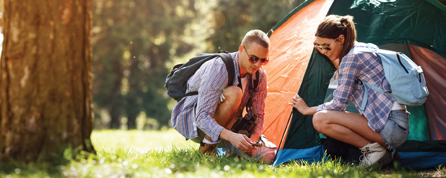 Two campers crouch beside a tent in a sunlit forest, working together to secure it. Both wear casual outdoor clothing and backpacks, with tall trees and soft morning light in the background.