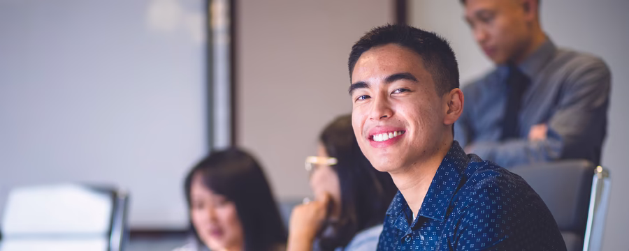 A young man in a blue patterned shirt smiles at the camera while sitting at a desk in a meeting room. Other people in the background are working on laptops and talking, suggesting a collaborative office or classroom setting.