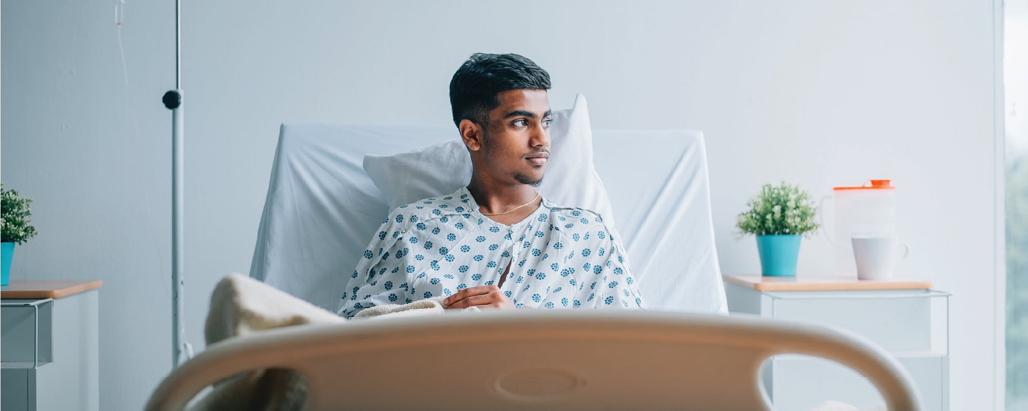 A hospital patient wearing a patterned gown sits upright in a hospital bed, looking thoughtfully toward a window. An IV bag hangs beside the bed, while a bedside table with a small potted plant and medical container adds to the clean, softly lit hospital room.