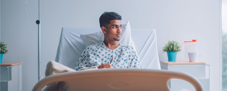 A hospital patient wearing a patterned gown sits upright in a hospital bed, looking thoughtfully toward a window. An IV bag hangs beside the bed, while a bedside table with a small potted plant and medical container adds to the clean, softly lit hospital room.