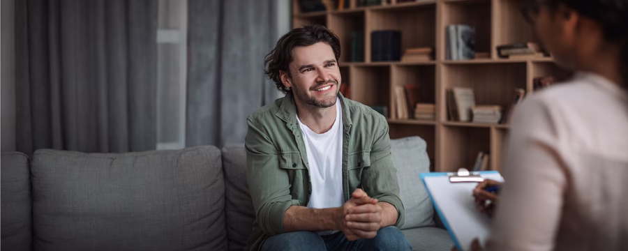A smiling man in a green shirt sits on a couch during a healthcare consultation with a provider in a white coat, who holds a clipboard. Warm wooden shelving lines the background of the clinical office setting.