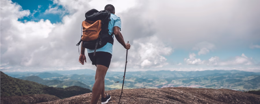 A hiker with a large backpack stands on a rocky mountaintop, holding a walking stick and looking out over a wide landscape of hills under a partly cloudy sky.