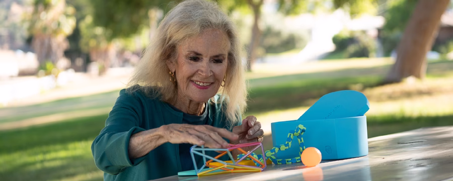 An older woman, Angela Fairhurst from Geri-Gadgets, with long gray hair smiles while sitting at a picnic table outdoors, reaching toward a colorful geometric toy and blue box in a sunny park.