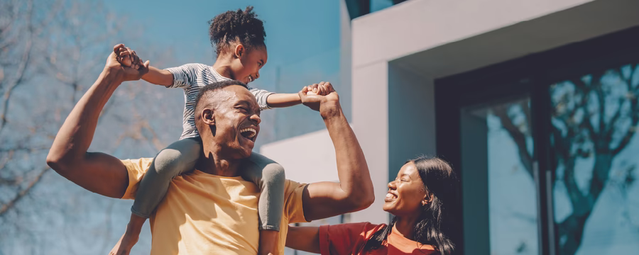 A joyful family outdoors on a sunny day, with a man carrying a young child on his shoulders while smiling, as a woman stands beside them laughing and reaching toward the child.