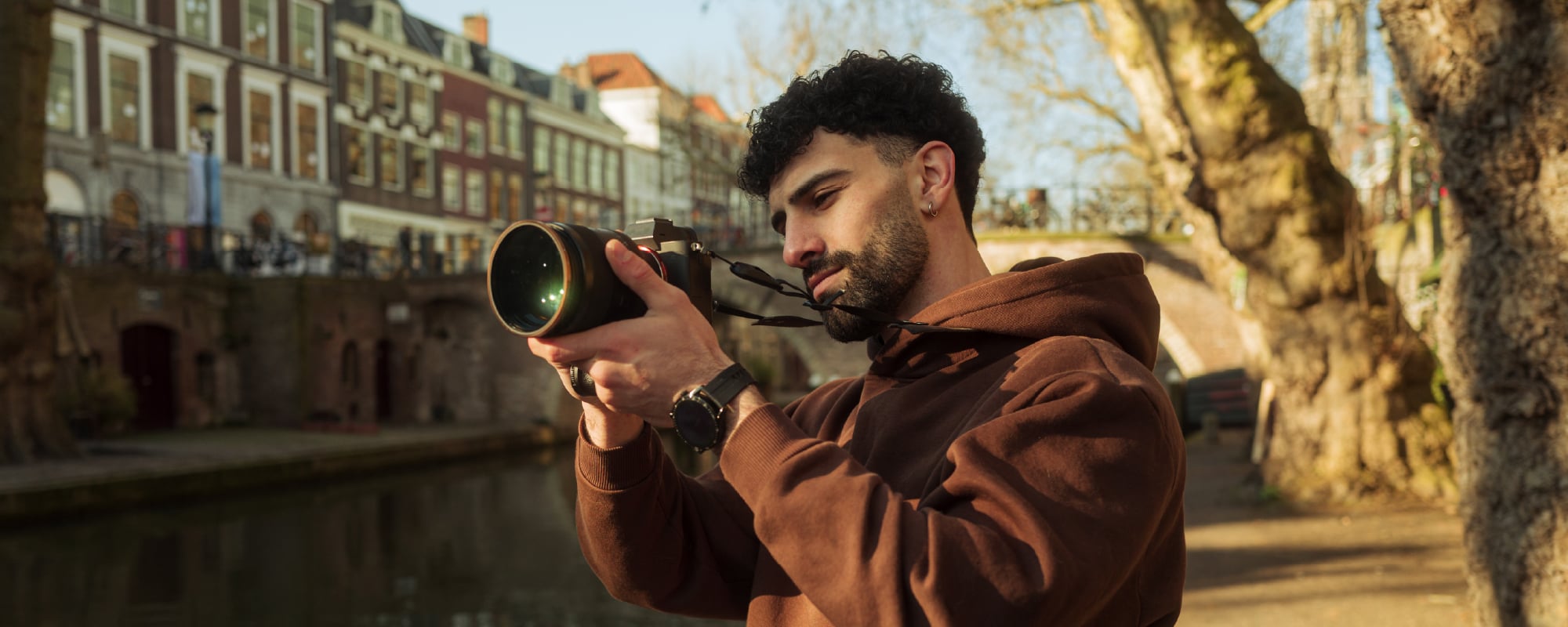 Person in a brown hoodie uses a mirrorless camera with a telephoto lens to take photos along a tree-lined canal in a historic city during golden hour.