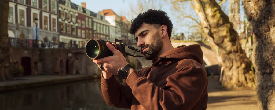 Person in a brown hoodie uses a mirrorless camera with a telephoto lens to take photos along a tree-lined canal in a historic city during golden hour.