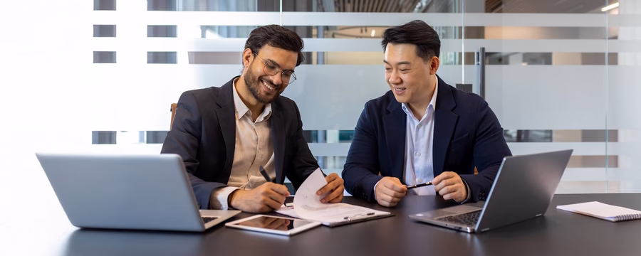 Two businessmen in a modern office setting, reviewing documents and working together at a table with laptops and a tablet.