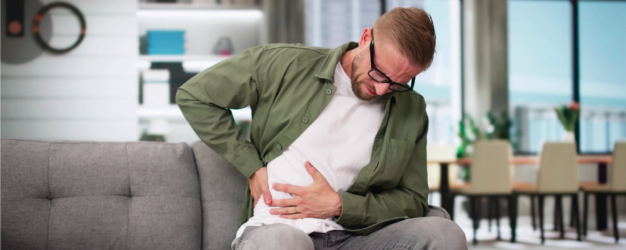 A man sitting on a couch grimaces in pain while clutching the lower right side of his abdomen with one hand and supporting his back with the other, suggesting abdominal or side pain in a home living room setting.