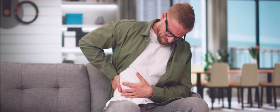 A man sitting on a couch grimaces in pain while clutching the lower right side of his abdomen with one hand and supporting his back with the other, suggesting abdominal or side pain in a home living room setting.