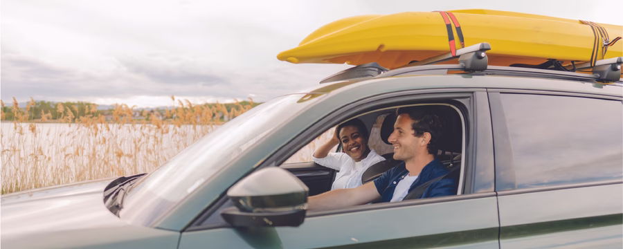 Two people smile from the front seats of a green SUV with two yellow kayaks secured to the roof rack, parked beside a lakeshore with tall reeds in the background.
