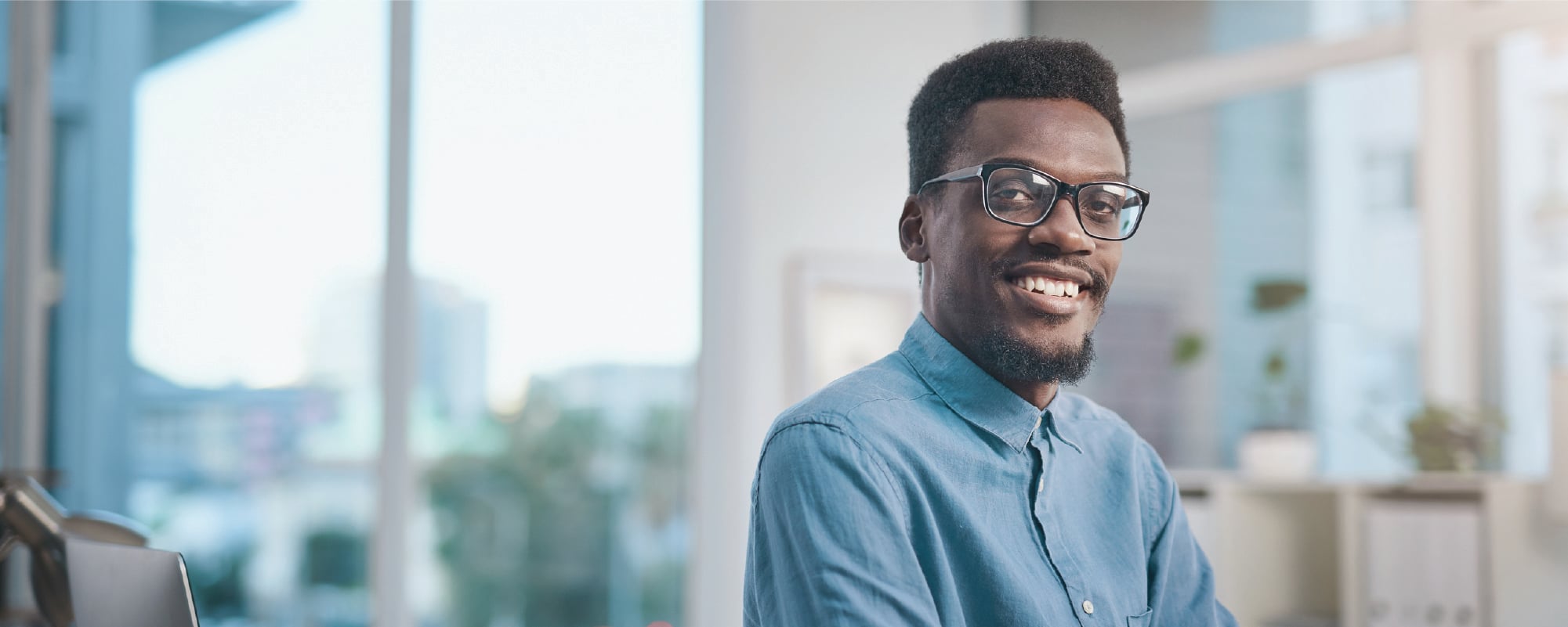 A smiling person wearing glasses and a light blue button-up shirt looks at the camera in a bright office setting. Large windows and blurred shelves in the background create a clean, professional atmosphere.