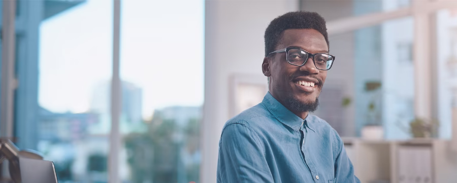 A smiling person wearing glasses and a light blue button-up shirt looks at the camera in a bright office setting. Large windows and blurred shelves in the background create a clean, professional atmosphere.