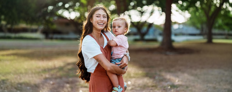 Smiling mother holding her young child outdoors in a sunlit park.