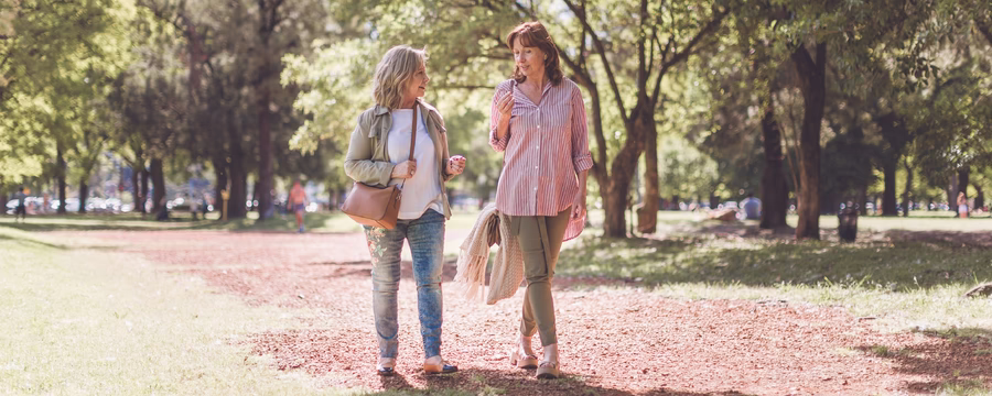 Two women walk side by side along a sunlit park path, smiling and talking while carrying light jackets. Tall trees cast dappled shade over the trail, creating a warm, relaxed outdoor setting.