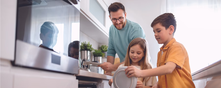 A man and two children load dishes into an open dishwasher in a bright, modern kitchen, smiling and working together.