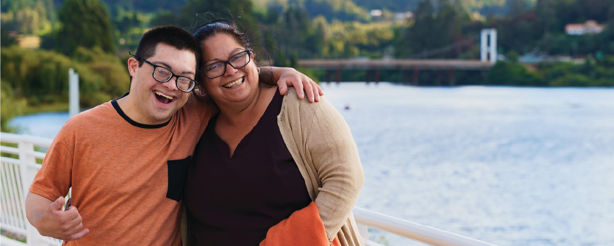 Two smiling people stand arm in arm on a lakeside walkway, posing for a photo with water, trees, and rolling hills in the background. One wears an orange T-shirt, while the other wears a black top with a beige cardigan and carries an orange jacket.