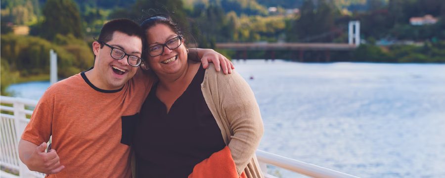 Two smiling people stand arm in arm on a lakeside walkway, posing for a photo with water, trees, and rolling hills in the background. One wears an orange T-shirt, while the other wears a black top with a beige cardigan and carries an orange jacket.